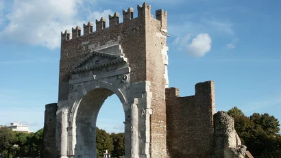 Arco romano in pietra bianca con mura di mattoni in un parco verde sotto cielo azzurro
